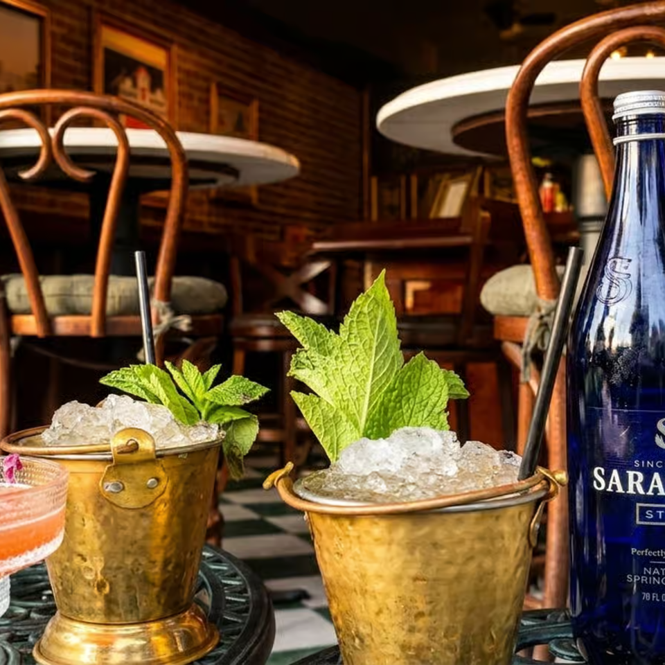 Cocktails garnished with mint and crushed ice sit beside Saratoga Still water bottles on a warmly lit restaurant patio.