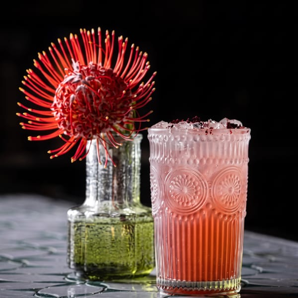 Vibrant red cocktail and a protea flower in a vase on a patterned, reflective surface.