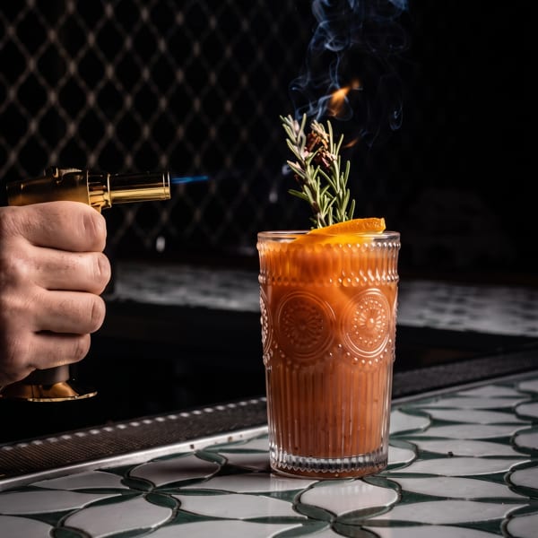 Hand torches a smoky orange cocktail garnished with rosemary on a patterned bar.