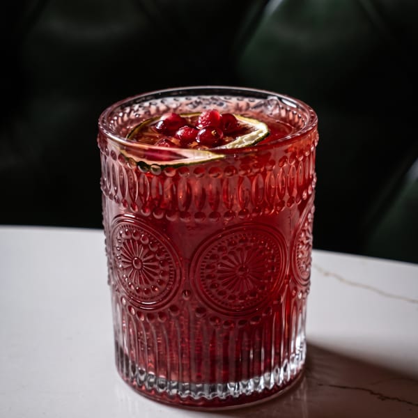 Red cocktail in an ornate glass, garnished with cranberries and a lime slice on a white marble table.