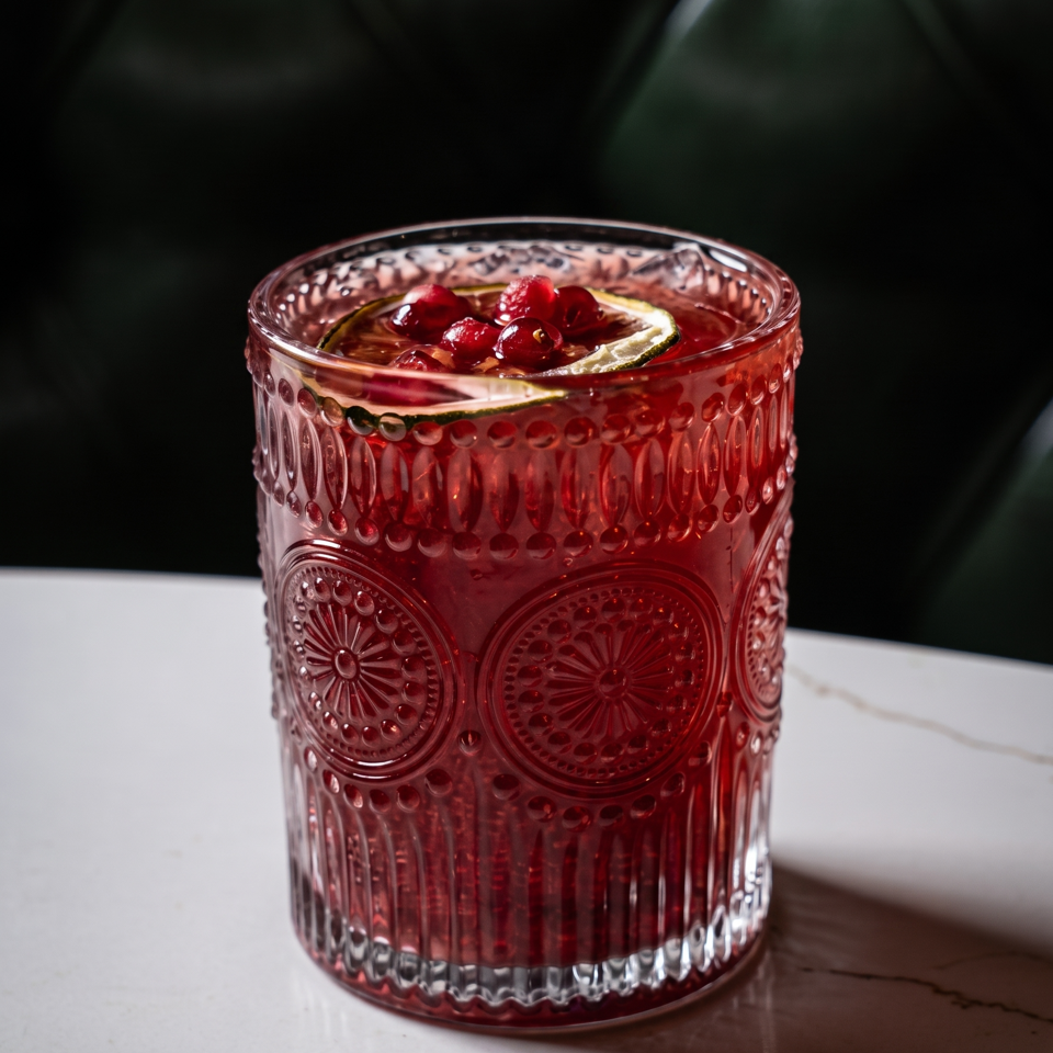 Red cocktail with berries and citrus garnish in an ornate textured glass on a marble table.