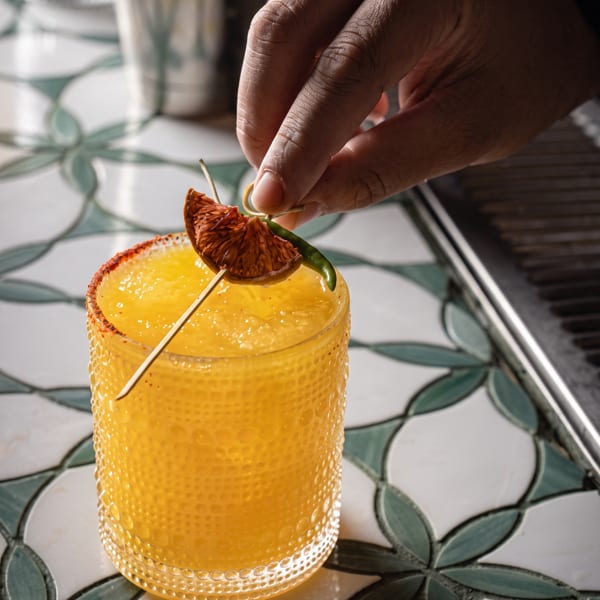 A hand garnishes a vibrant orange cocktail with a dried orange slice and green chili.