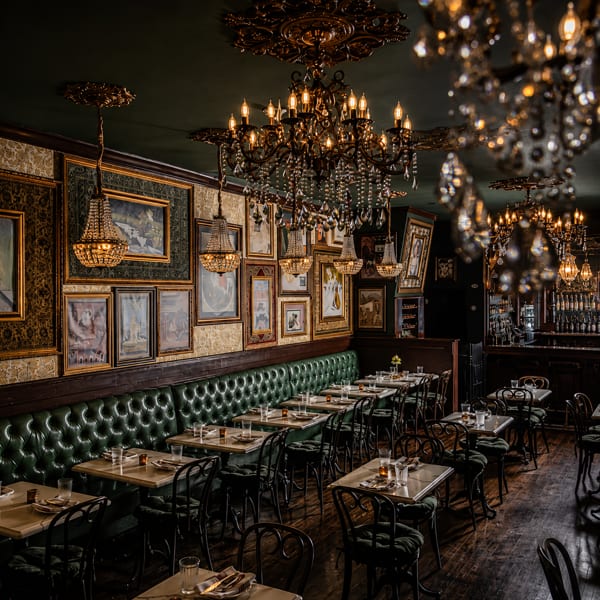 Luxurious restaurant dining room with dark green banquettes, ornate chandeliers, and framed artwork on patterned walls.